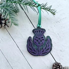 A decorative thistle-shaped ornament in dark green and purple, hanging from a green ribbon on a white wooden surface with pine branches and pinecones around it.