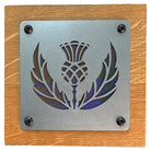 A square metal plaque featuring a laser-cut thistle and laurel design, mounted on a square wooden board with four visible black screws.