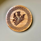 A round wooden wall plaque featuring an engraved Celtic knot border and a central thistle design, mounted on a light beige wall.