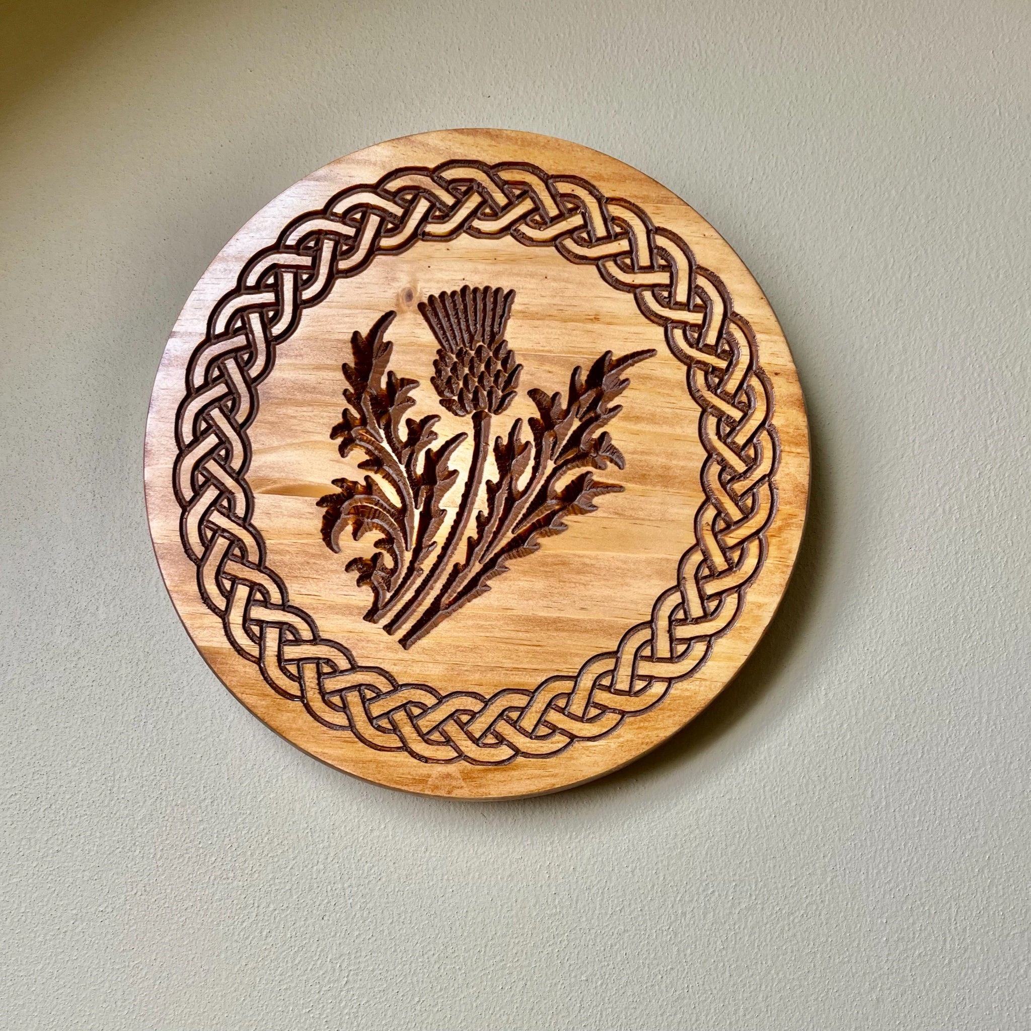 A round wooden wall plaque featuring an engraved Celtic knot border and a central thistle design, mounted on a light beige wall.