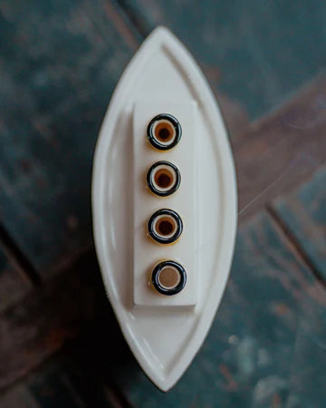 A white ceramic boat-shaped incense holder with four vertical slots containing small black and gold beads, photographed from above on a dark rustic wooden surface.