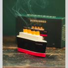 A Titanic-themed smoking accessory shaped like the ship, with visible smoke rising from its yellow funnels, placed in front of a dark branded box on a rustic wooden surface.