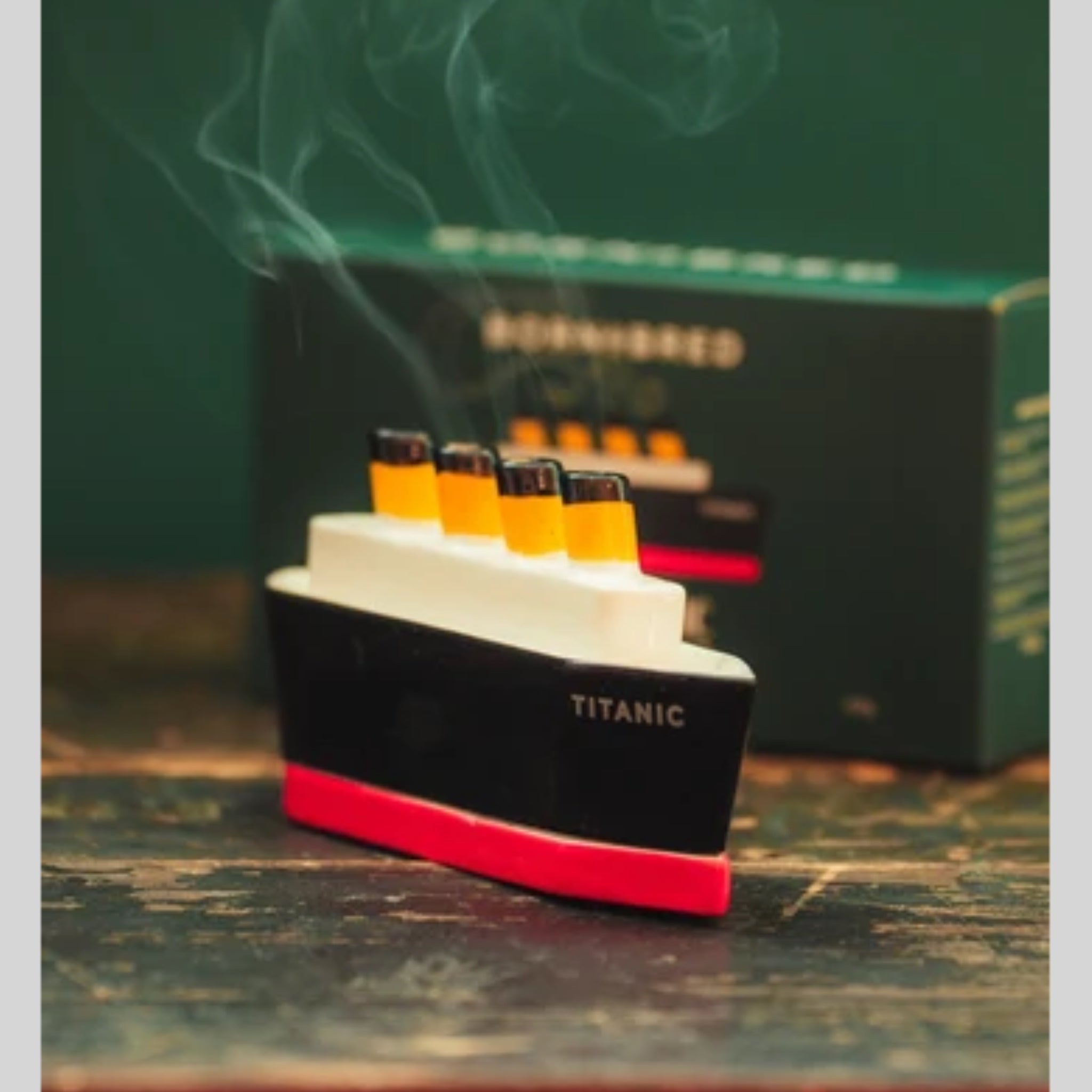 A Titanic-themed smoking accessory shaped like the ship, with visible smoke rising from its yellow funnels, placed in front of a dark branded box on a rustic wooden surface.