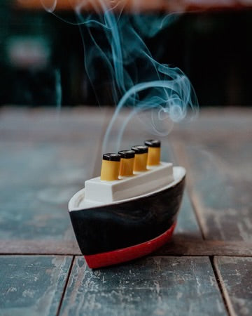 A miniature model ship incense holder with four lit yellow fountains emitting blue smoke, set on a rustic wooden surface.