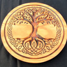 A round wooden plaque featuring a carved Celtic-style tree of life with roots and branches integrated into an interlaced knot border, shown on a black background.