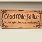 A rectangular wooden sign with Celtic knot borders and the carved text 'Céad Mile Féilte' and '“A hundred thousand welcomes” on a light beige background.