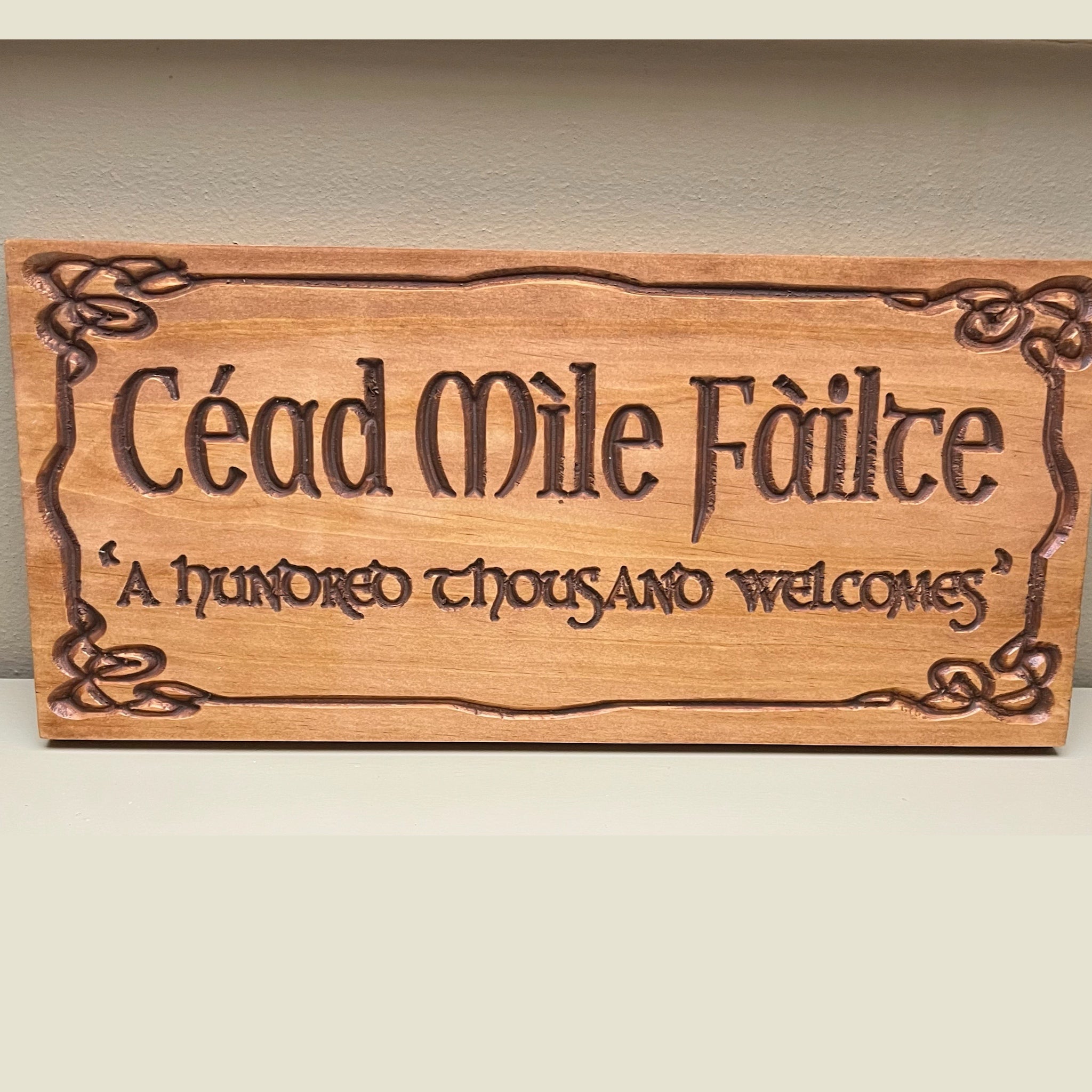 A rectangular wooden sign with Celtic knot borders and the carved text 'Céad Mile Féilte' and '“A hundred thousand welcomes” on a light beige background.