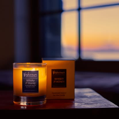 A lit 'Whisky & Honey' scented candle in a clear glass jar sits on a dark wooden table next to its yellow packaging box, with a warm sunset window in the background.