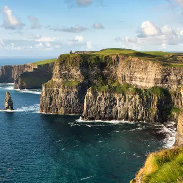 A scenic coastal landscape with tall rocky cliffs topped with green grass, overlooking deep blue ocean water with white surf and a partly cloudy sky.