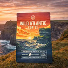 A bag of Wild Atlantic Coffee stands upright on grassy ground with a dramatic coastal cliff and ocean sunset in the background. The package features an illustrated seascape with a red-orange sky, waves, and dark blue lower section.