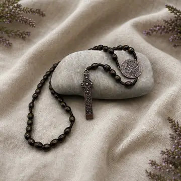 A dark brown beaded rosary with a silver-toned crucifix and medallion, draped over a smooth light gray stone on a beige linen fabric background with dried lavender sprigs.
