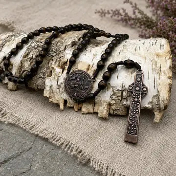 A dark wooden bead rosary with a round medallion and a cross pendant, styled on a piece of birch wood over a burlap fabric background.