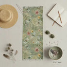 A rectangular scarf laid flat on a light neutral surface, surrounded by styled props including a straw hat, sunglasses, a notebook, dried stems, and a small bowl. The scarf has a soft sage green base with a delicate floral print in muted pink, cream, and yellow tones.