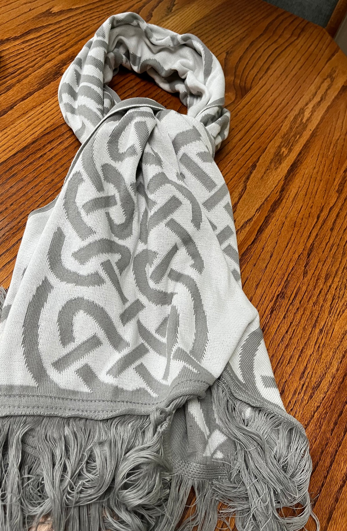 A light gray and white knit scarf with a geometric interlocking pattern and fringed ends, laid out on a wooden table.